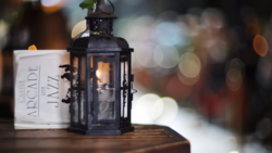 Black metal lantern with candle flame beside a white candle container, shot with shallow depth of field and warm bokeh background.