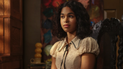 Woman with long dark curly hair wearing a cream-colored blouse with puff sleeves and tie neckline, looking off-camera in an interior setting.