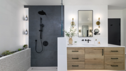 Modern bathroom interior with dark tile shower enclosure, wooden vanity, and brass-accented mirrors and lighting fixtures.