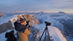 Photographer on snowy alpine ridge with camera on tripod overlooking snow-covered peaks at golden hour.