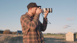 Photographer in plaid shirt and cap using telephoto lens outdoors at golden hour.
