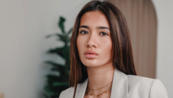 Woman in cream blazer with long dark hair against neutral interior background.