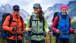 Three female hikers with backpacks and trekking poles posing on a mountain trail with alpine peaks in the background.
