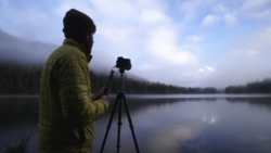 Photographer in yellow jacket operating tripod-mounted camera beside misty lake with forested shoreline.