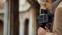 Photographer with white hair holding a large format film camera to their eye in an urban courtyard setting.