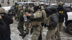 Armed personnel in tactical gear and winter clothing gathered in a snowy urban plaza during daytime.