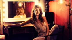 Woman in light-colored dress seated at illuminated vanity mirror in warm-toned studio setting.