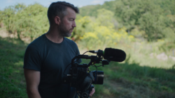 Videographer holding a professional cinema camera with shotgun microphone in an outdoor natural setting.