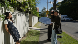 Photographer holding large reflector while shooting portrait of woman against white fence in natural daylight.