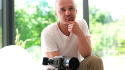 Male photographer holding a professional video camera while posing indoors near large windows with natural landscape visible outside.