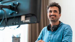Man in denim shirt smiling at camera in a studio setup with monitor and headphones.