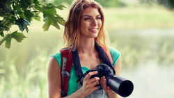 Woman holding binoculars outdoors near tree branches and blurred water background.