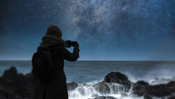 Silhouetted figure photographing the Milky Way above turbulent ocean waves at night.