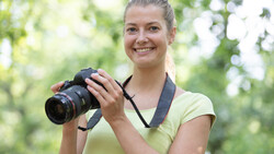 Young woman holding a DSLR camera with telephoto lens in outdoor setting with natural green foliage background.