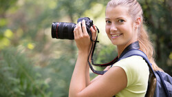Female photographer holding a DSLR camera with telephoto lens, smiling at camera in outdoor forest setting.