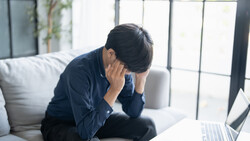 Man in dark blue shirt holding his head while sitting at laptop, expressing stress or fatigue.