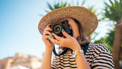 Photographer in striped shirt and straw hat holding vintage camera up to face against tropical backdrop.