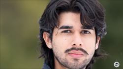Portrait of a man with shoulder-length dark hair and facial hair against a soft green background.