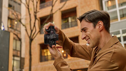 Photographer holding a vintage film camera up to his face, smiling while framing a shot in an urban setting.