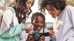 Three women smiling while looking at a camera display in outdoor daylight.