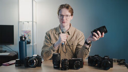 Man holding a camera lens while discussing photography gear displayed on a table.