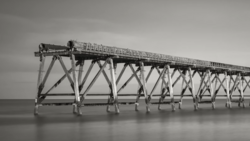 Long wooden pier with repetitive X-braced support structure extending into calm water under overcast sky.