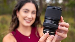 Woman holding a telephoto lens toward the camera in an outdoor garden setting.