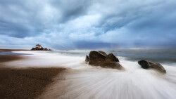 Long-exposure seascape with smooth water flowing around dark rocks on a sandy beach under stormy clouds.