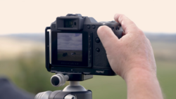 Photographer holding a medium format camera on a tripod, viewing the outdoor landscape through the LCD screen.