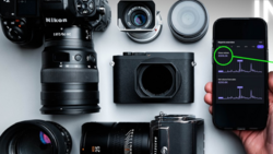 Flat lay of camera gear including a Nikon DSLR, multiple lenses, a mirrorless camera body, and a smartphone displaying histogram data.