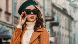 Woman wearing dark beret, sunglasses, and rust-colored blazer adjusting her sunglasses on urban street.