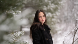 Woman with long dark hair wearing a black coat in a snowy forest setting.