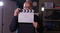 Bald man wearing glasses holds a film clapperboard marked "GARCON SOM" in a home studio setting.