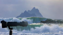Telephoto lens on tripod focused on large ocean waves with rocky mountain peak in background.