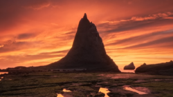 Dramatic silhouette of a tall sea stack against a vibrant orange and yellow sunset sky with reflection in wet tidal flats.