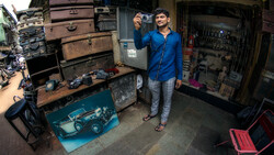 Man in blue jacket holding a camera in a cluttered workshop filled with vintage suitcases and tools.