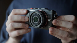 Hands holding a compact mirrorless camera with a fixed prime lens against a blue shirt.