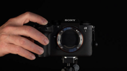 Hands holding a Sony Alpha mirrorless camera body without a lens attached, mounted on a tripod against a black background.