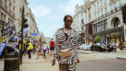 Woman in zebra-striped outfit walking on busy urban street lined with classical buildings.