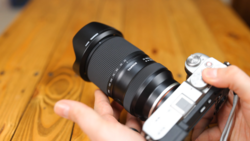 Hands holding a mirrorless camera with a telephoto lens mounted on a wooden surface.