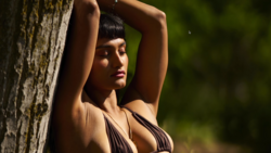 Woman leaning against a textured tree trunk, arms raised, gazing to the side in natural outdoor lighting.