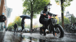 Man riding a motorcycle through a wet urban street during rain, splashing through water.