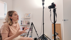 Woman sitting in a home studio setup, gesturing while speaking to camera during a recording session.