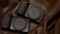 Two compact digital cameras with black bodies and textured grips positioned on brown leather fabric.