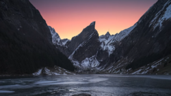 Snow-covered mountains frame a frozen alpine lake at dawn, with pink and orange sky reflected in still water.