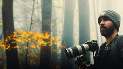 Bearded photographer in knit beanie holding telephoto lens in misty forest with golden foliage