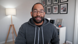 Man wearing glasses and dark hoodie smiling at camera in home office setting.