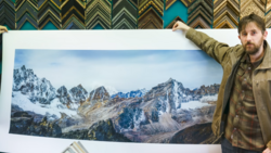 Photographer holding a large alpine landscape print featuring snow-capped peaks and rocky terrain.