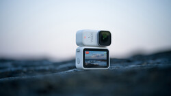 White action camera with flip-out screen displaying ocean footage, positioned on wet rocks with blurred seascape background.