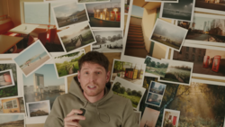 Man in tan hoodie gesturing toward a wall covered with dozens of printed photographs arranged in an overlapping collage.